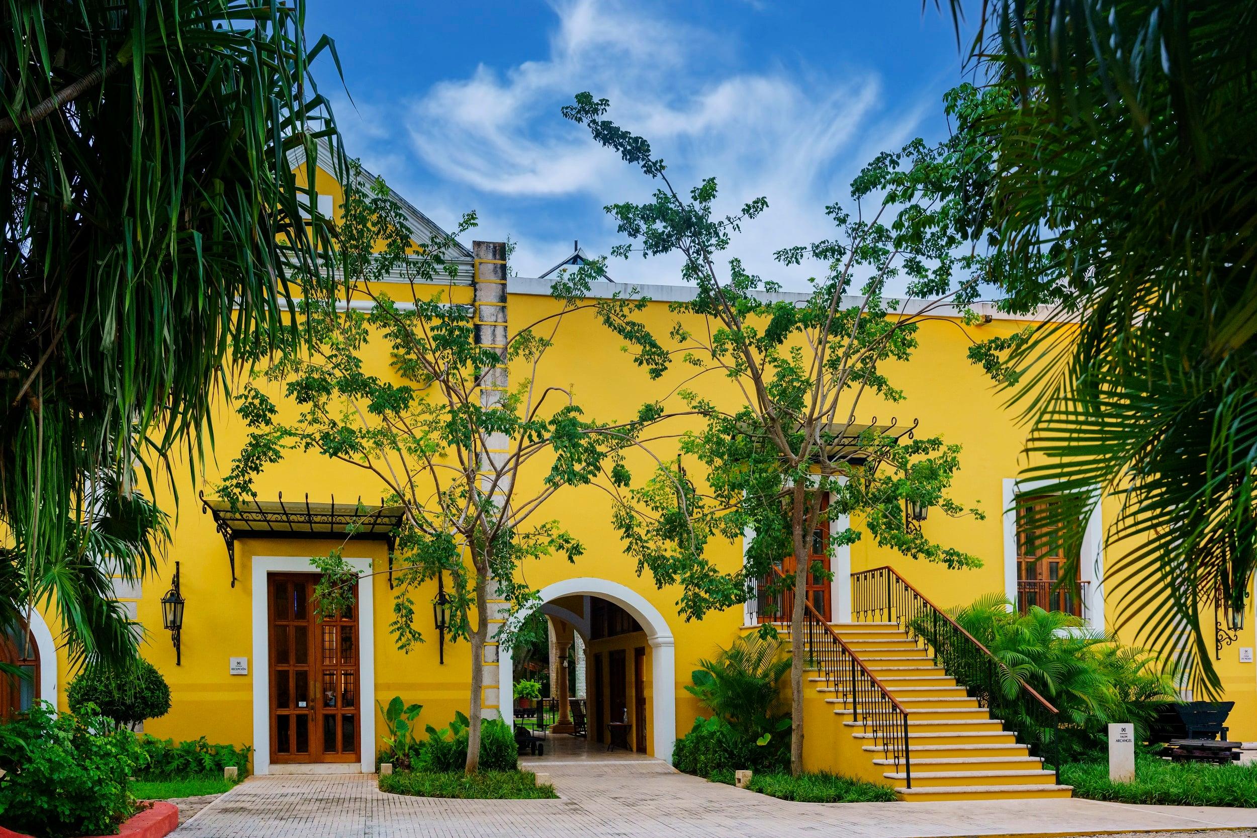 Bright yellow building with stairs surrounded by lush greenery at Hacienda Xcanatun, Angsana Heritage Collection.