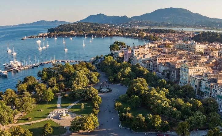 Aerial view shows Corfu coastline with lush trees, a marina of sailboats, and cityscape near Angsana Corfu Resort & Spa.