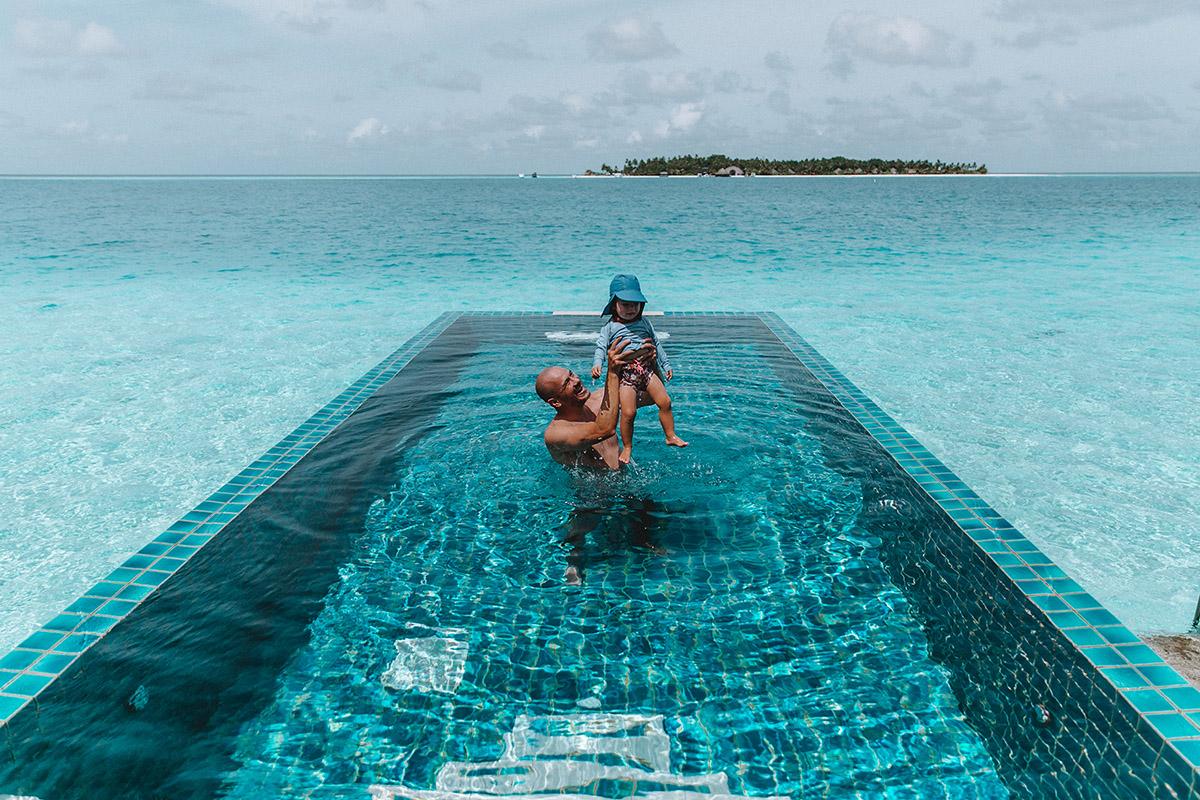 Dad playfully holding child in crystal-clear pool at Angsana Velavaru Maldives.