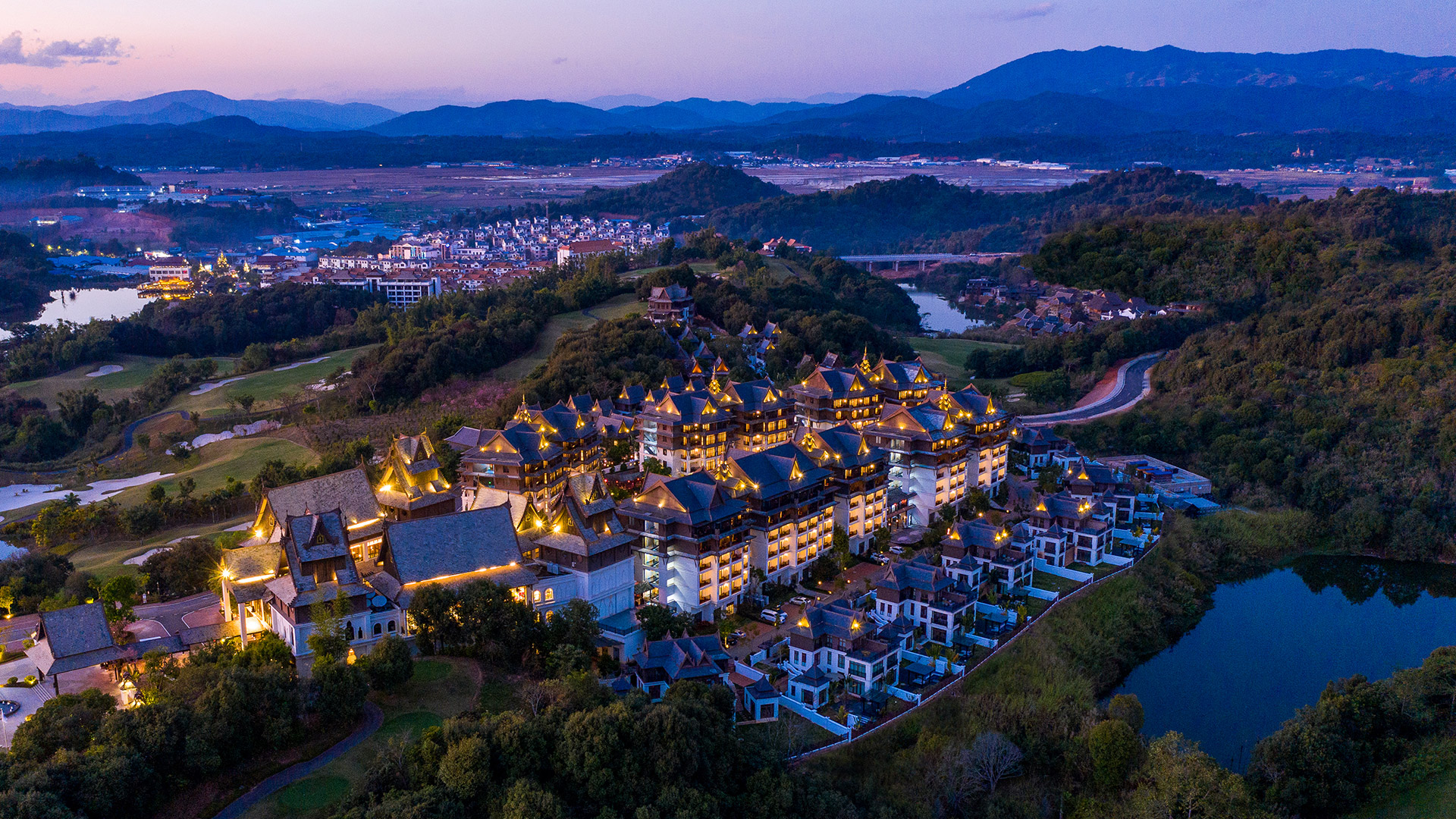 Angsana Xishuangbanna resort illuminated at dusk, nestled in lush hills with distant mountains.
