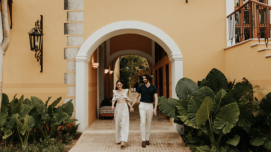 Couple walking hand-in-hand under Hacienda Xcanatun, Angsana Heritage Collection's archway amidst lush greenery.