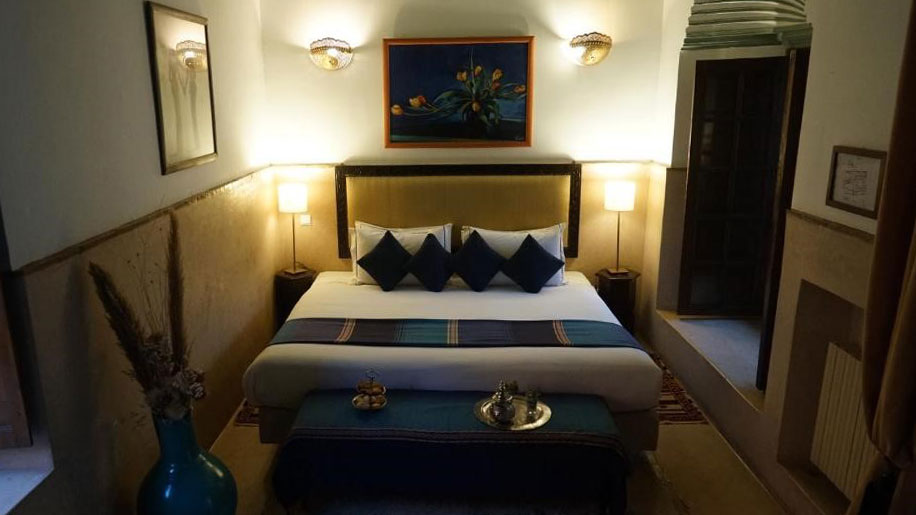 A cozy room features a bed with blue accents, softly lit lamps, and elegant decor at Angsana Riads Collection - Marrakech, Si Said Deluxe King.