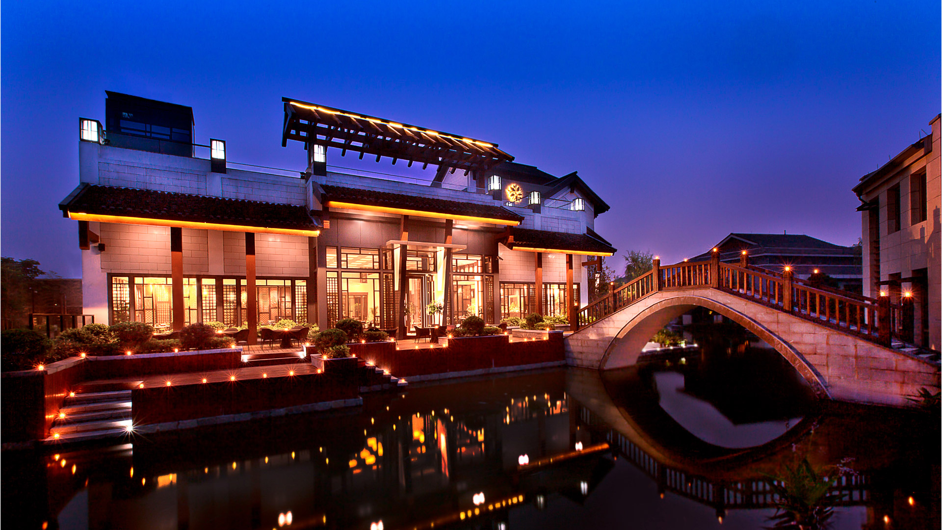 Illuminated building reflects in water, beside a lit bridge at Angsana Hangzhou China.