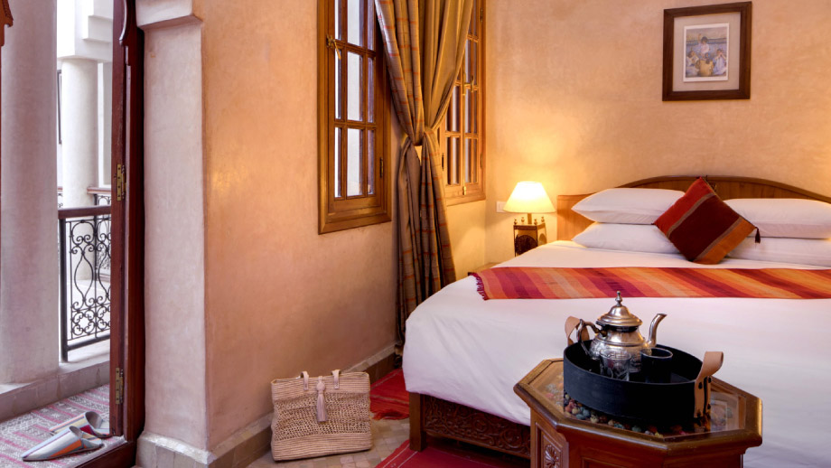 Cozy bed with colorful pillows; tea tray beside; open balcony door in Angsana Riads Collection - Marrakech, Lydines Studio King.