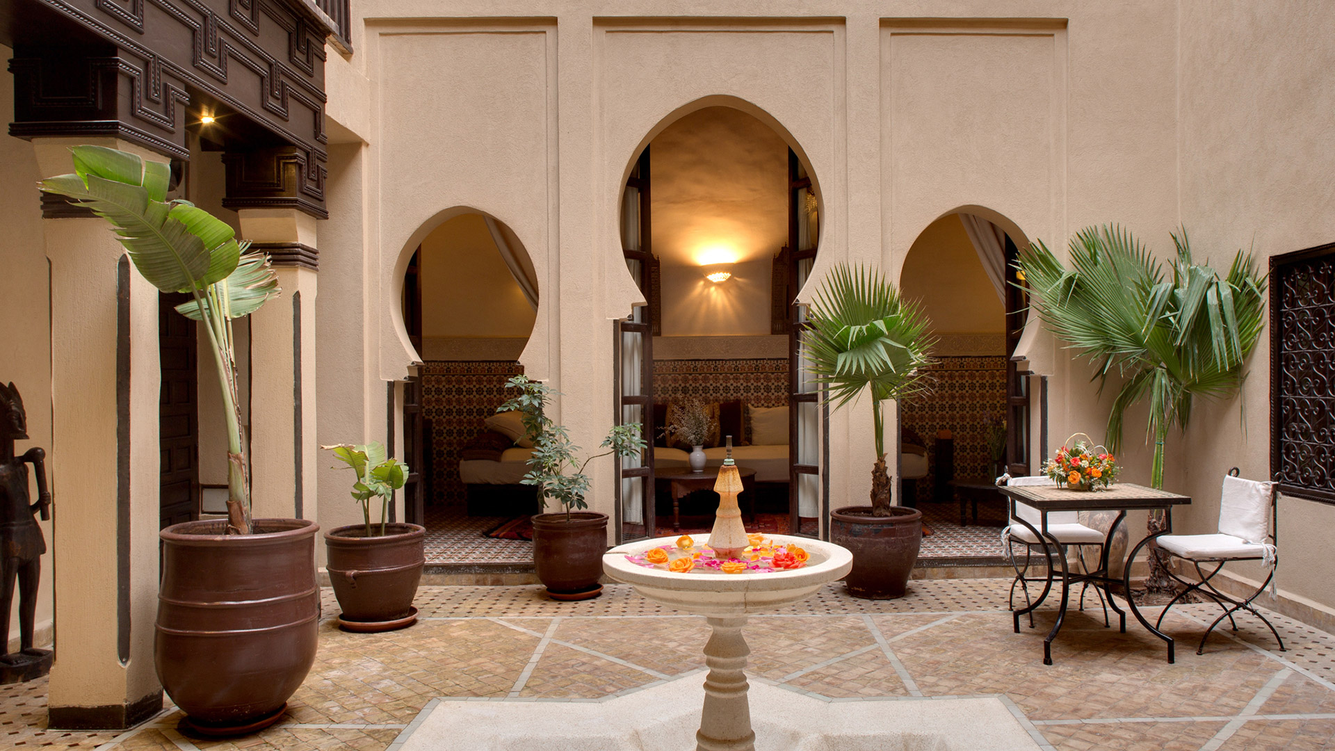 Fountain with floating petals stands in a serene courtyard, framed by arches and potted plants at Angsana Riads Collection - Marrakech, Riad Tiwaline.