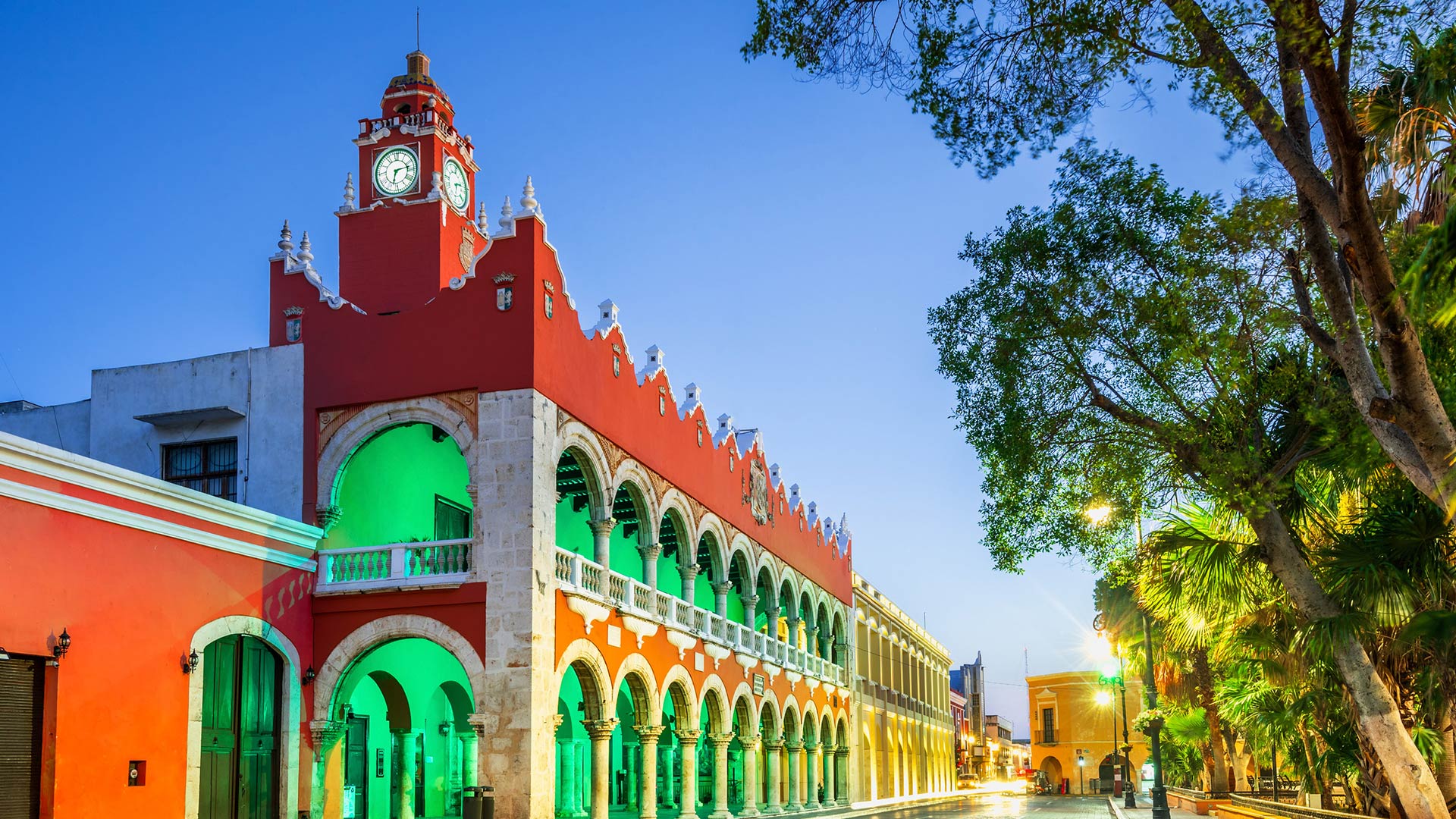 Clock tower glows green, framed by vibrant trees at Hacienda Xcanatun, Angsana Heritage Collection.