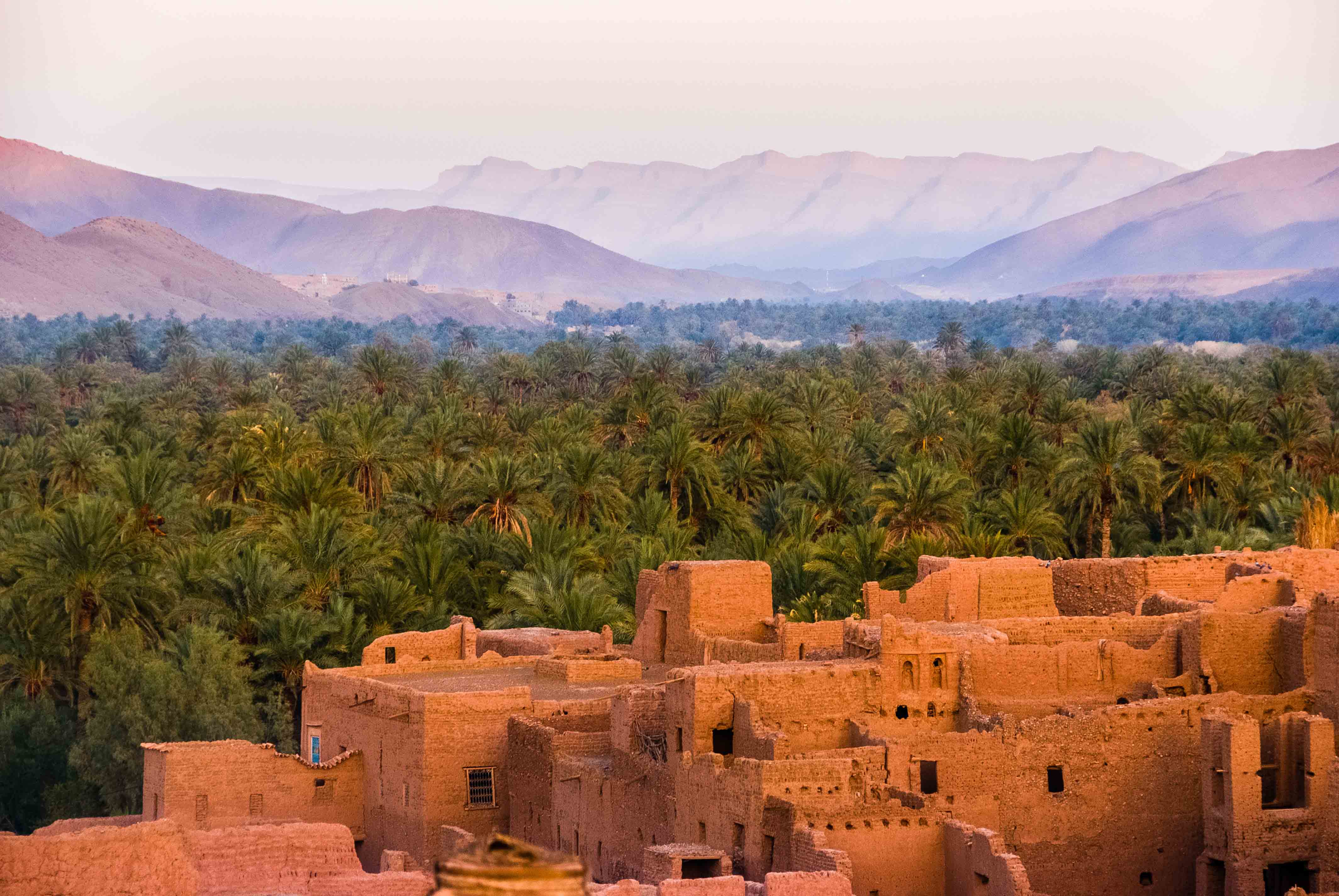 Ancient brick buildings stand amidst lush palm trees and distant mountains at Angsana Riads Collection - Marrakech.