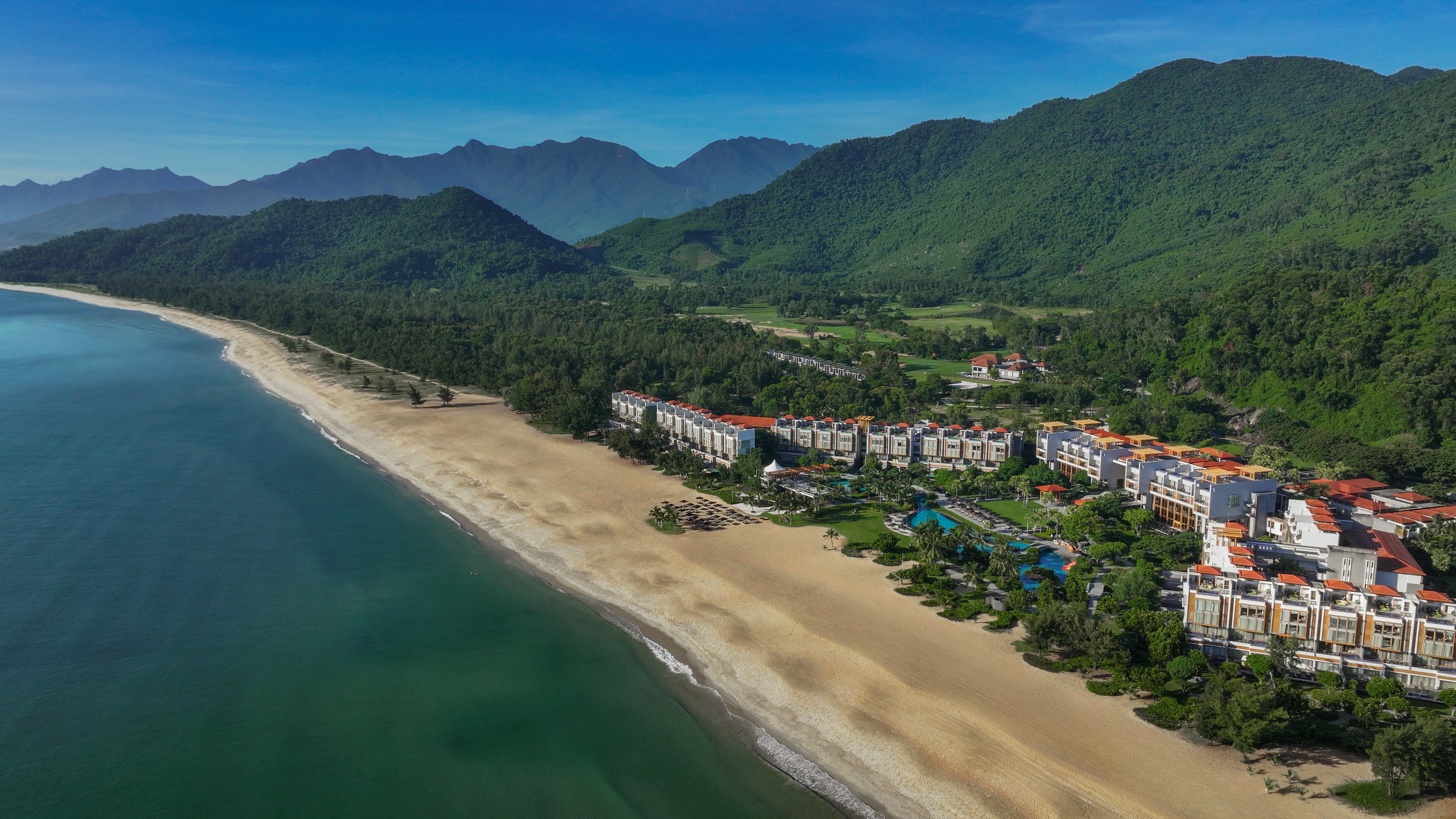 Angsana Lăng Cô resort spans a sandy beach, bordered by lush green mountains.