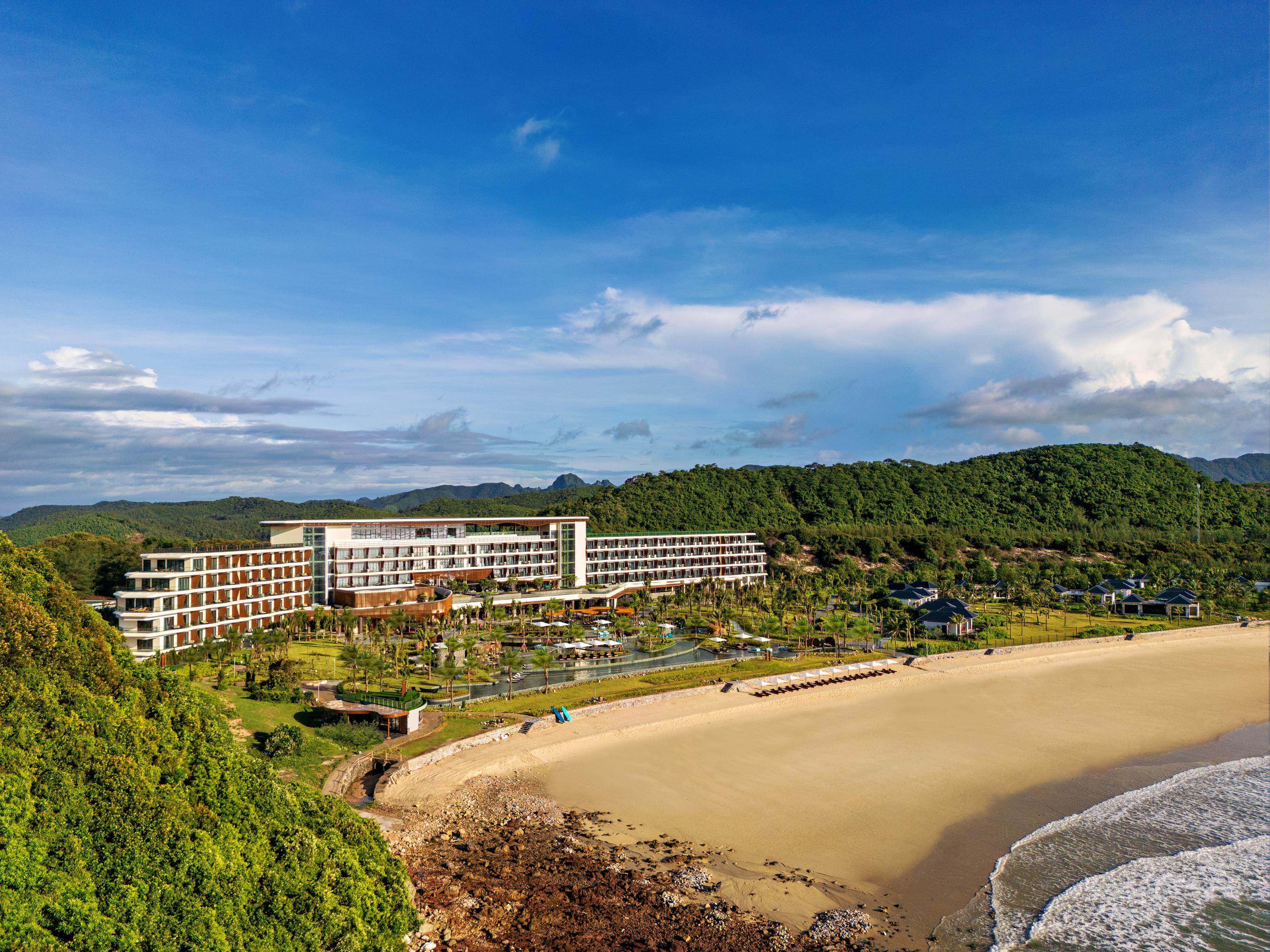 Hotel Angsana Quan Lan overlooks a beach, surrounded by lush green hills and clear skies.